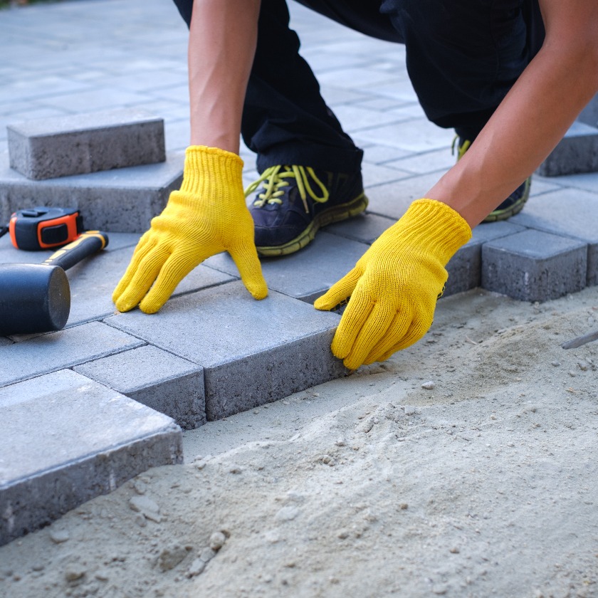 The master in yellow gloves lays paving stones in layers. Garden brick pathway paving by professional paver worker. Laying gray concrete paving slabs in house courtyard on sand foundation base.